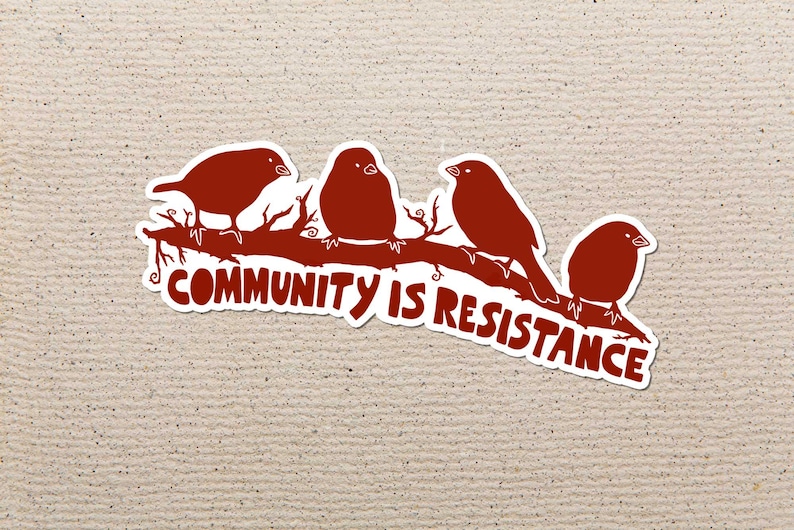 May include: A sticker featuring four red birds perched on a brown branch. The text "COMMUNITY IS RESISTANCE" is written in white below the birds. The sticker has a white border and is set against a textured beige background.