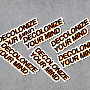 Könnte beinhalten: Eine Ansammlung weißer Aufkleber mit dem Text "DECOLONIZE YOUR MIND" in schwarzer und orangefarbener Farbverlaufsschrift. Die Aufkleber sind auf einer grauen Oberfläche angeordnet.