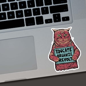 May include: A sticker featuring a red cat with green eyes holding a sign that reads "EDUCATE ORGANIZE REVOLT." The cat is on a laptop, and the sticker has a distressed, hand-drawn style.