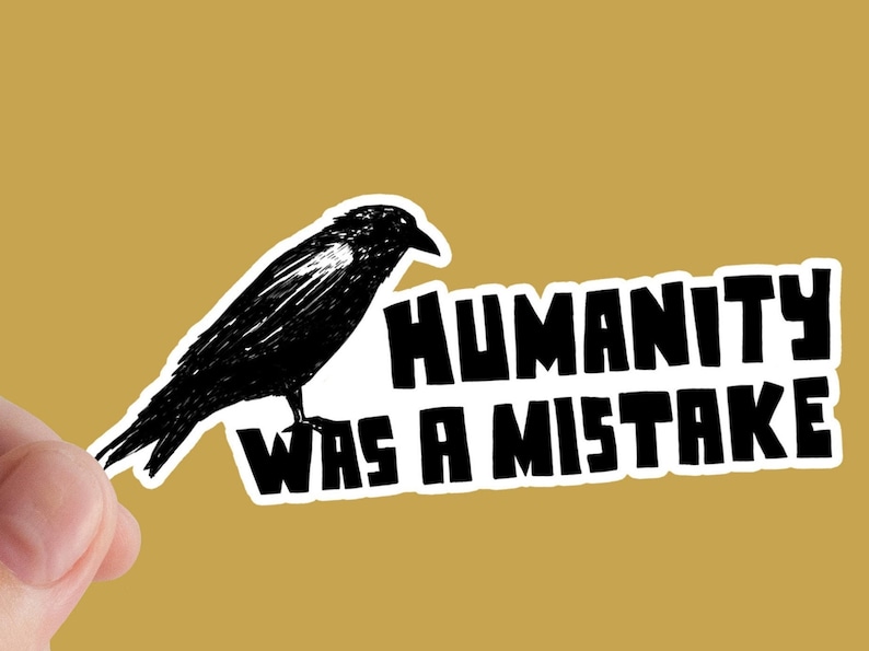 May include: A black and white sticker featuring a raven illustration and the text "HUMANITY WAS A MISTAKE". The sticker is held against a gold background.