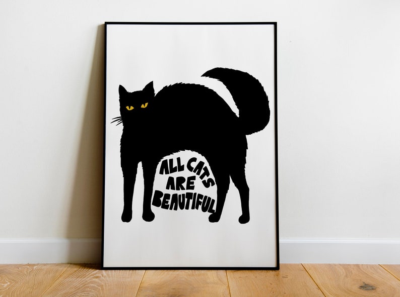 May include: Black cat silhouette art print with yellow eyes and the text "ALL CATS ARE BEAUTIFUL" on a white background. The artwork is framed in a black frame and displayed on a wooden floor.