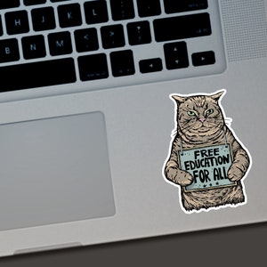 Puede incluir: Una pegatina con un gato gru&ntilde;&oacute;n sosteniendo un cartel que dice "FREE EDUCATION FOR ALL". El gato es de color marr&oacute;n claro y gris con ojos verdes. El cartel es azul claro con texto blanco y estrellas.
