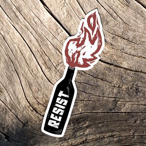 May include: A sticker featuring a black bottle with a red flame design and the word "RESIST" in white. The sticker is placed on a weathered, light brown wooden surface.