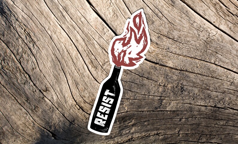 May include: A sticker featuring a black bottle with a red flame design and the word "RESIST" in white. The sticker is placed on a weathered, light brown wooden surface.