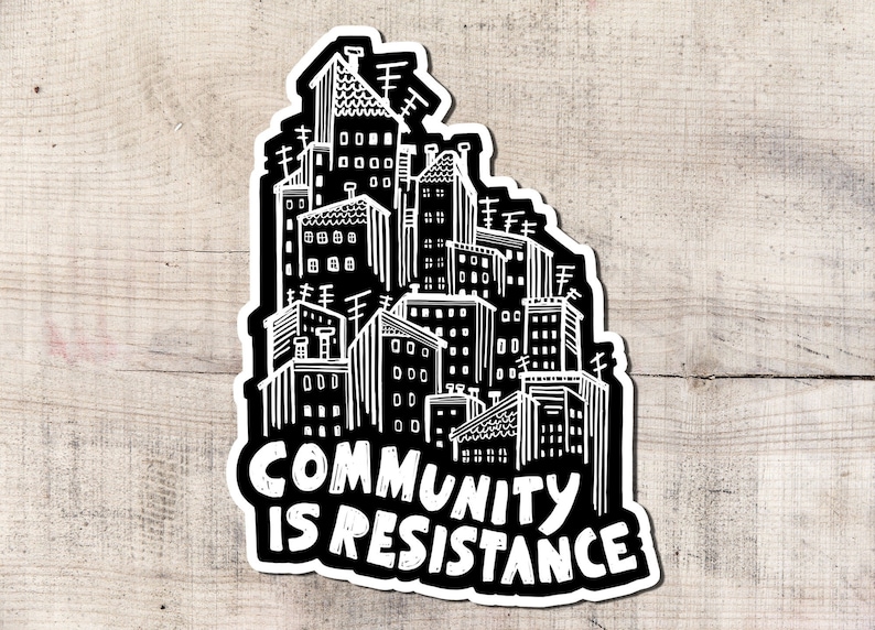 May include: A black and white sticker featuring a stylized illustration of a cityscape. The buildings are densely packed, with various architectural details. The text "COMMUNITY IS RESISTANCE" is prominently displayed in bold, white lettering.
