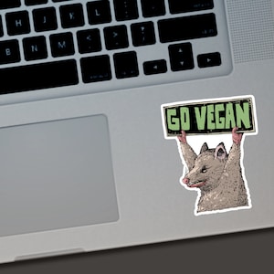 May include: A sticker featuring a cartoon opossum holding a sign that reads "GO VEGAN." The opossum is depicted in shades of brown, gray, and pink, with a determined expression. The sign has a black border and green text.