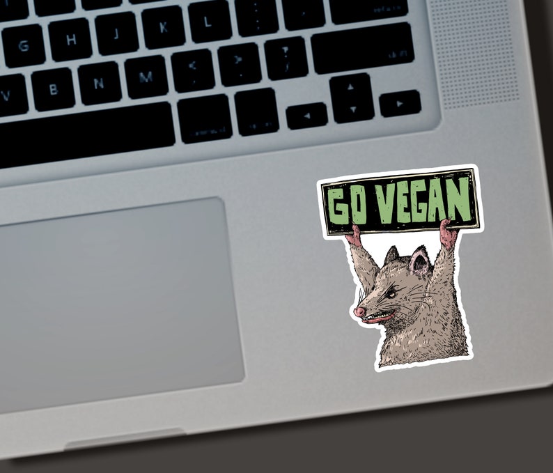 May include: A sticker featuring a cartoon opossum holding a sign that reads "GO VEGAN." The opossum is depicted in shades of brown, gray, and pink, with a determined expression. The sign has a black border and green text.