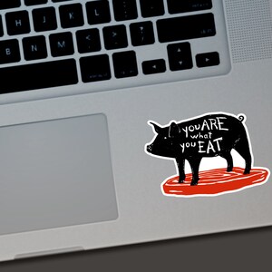 May include: A black sticker featuring a pig silhouette with the text "you are what you eat" in white. The pig stands on a red, steak-shaped base. The sticker is placed on a laptop.