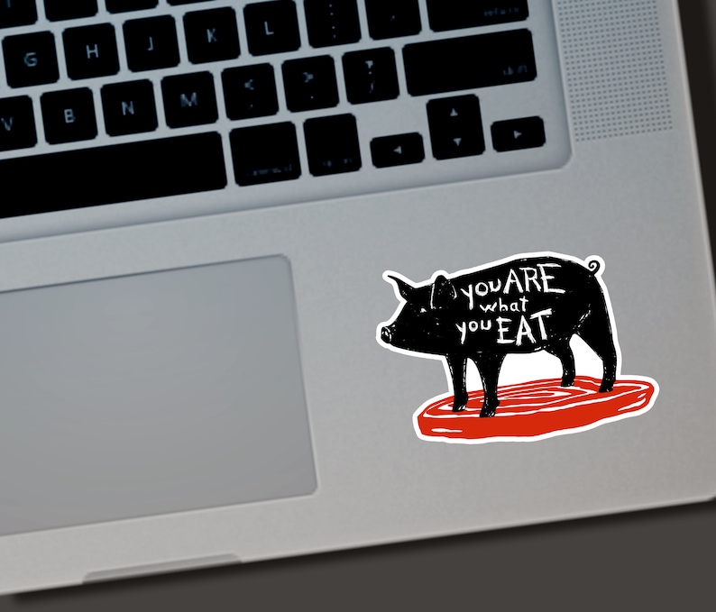 May include: A black sticker featuring a pig silhouette with the text "you are what you eat" in white. The pig stands on a red, steak-shaped base. The sticker is placed on a laptop.