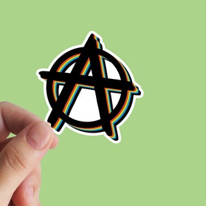 Puede incluir: Pegatina con el s&iacute;mbolo de la anarqu&iacute;a en negro, con contorno de arco&iacute;ris y borde blanco. El s&iacute;mbolo es una letra "A" dentro de un c&iacute;rculo. La pegatina est&aacute; sujeta por una mano sobre un fondo verde claro.