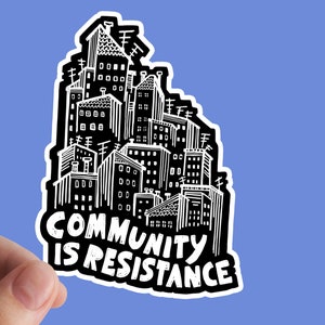 May include: A black and white sticker featuring a cityscape illustration. The buildings are densely packed with detailed windows, chimneys, and antennas. Bold text reads "COMMUNITY IS RESISTANCE" in a stylized font, set against a blue background.