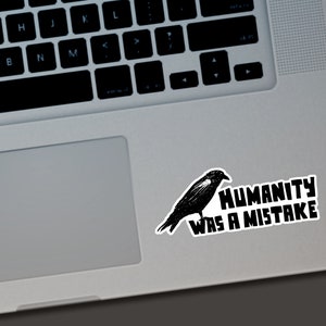 May include: A black sticker featuring a detailed illustration of a crow perched on a branch. The sticker has the text "HUMANITY WAS A MISTAKE" in bold, white letters. The sticker is placed on a laptop.
