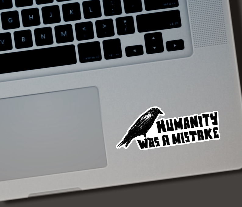May include: A black sticker featuring a detailed illustration of a crow perched on a branch. The sticker has the text "HUMANITY WAS A MISTAKE" in bold, white letters. The sticker is placed on a laptop.