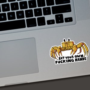 May include: A sticker featuring a cartoon crab in shades of yellow and brown. The crab has large claws and is positioned above the text "EAT YOUR OWN FUCKING HANDS" in a bold, black font. The sticker is on a laptop.