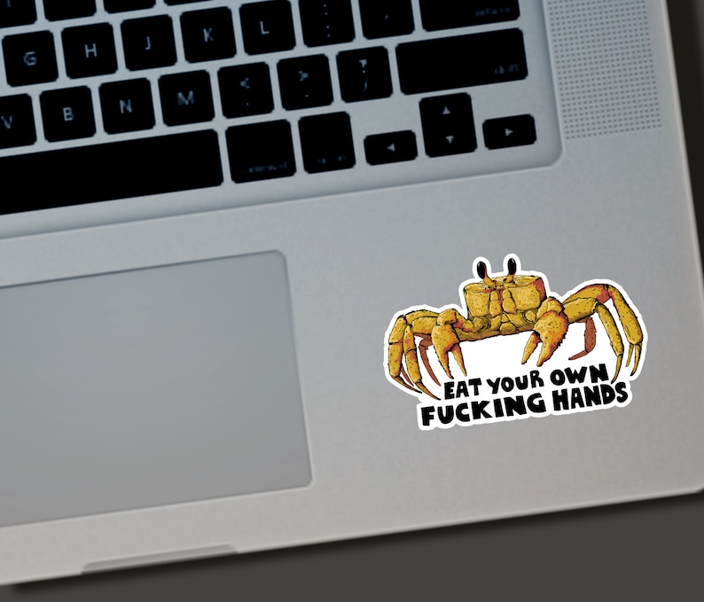 May include: A sticker featuring a cartoon crab in shades of yellow and brown. The crab has large claws and is positioned above the text "EAT YOUR OWN FUCKING HANDS" in a bold, black font. The sticker is on a laptop.