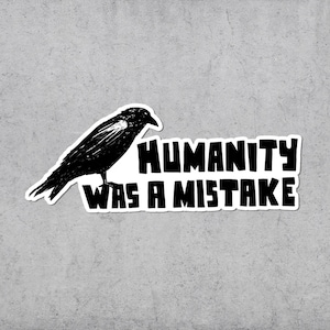 May include: A black and white sticker featuring a detailed illustration of a crow perched above the text "HUMANITY WAS A MISTAKE" in bold, block letters. The sticker has a white border and is set against a gray background.