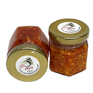 May include: Two small glass jars filled with a red and orange chili pepper paste. The jars have gold lids with labels that say "100% Natural Paste 1.5 oz. www.charapitafarms.com"