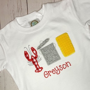 May include: A white short-sleeved t-shirt featuring an embroidered design of a red and white plaid lobster, a gray trash can, and a yellow rectangular sponge. The name Greyson is embroidered in red cursive text below the graphic elements.