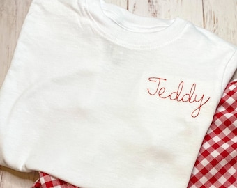 Puede incluir: Camiseta blanca con el nombre "Teddy" bordado en rojo en la parte delantera, combinada con pantalones cortos de cuadros rojos y blancos.