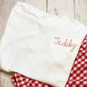 May include: White t-shirt with the name "Teddy" embroidered in red on the front, paired with red and white gingham shorts.