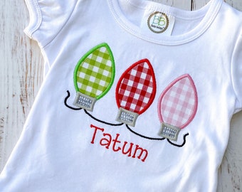 Puede incluir: Camiseta blanca de manga corta con un dobladillo con volantes. La camiseta presenta un diseño bordado de tres luces de Navidad en patrones de cuadros verdes, rojos y rosas. El nombre "Tatum" está bordado debajo de las luces.