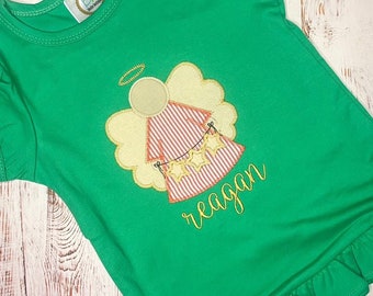 Puede incluir: Una camiseta verde brillante de manga corta con dobladillo con volantes y una aplicación bordada central de un ángel. El ángel tiene un vestido de rayas rojas y blancas, alas amarillas, un halo y sostiene una cadena de estrellas amarillas. El nombre Reagan está bordado en cursiva amarilla debajo del diseño.