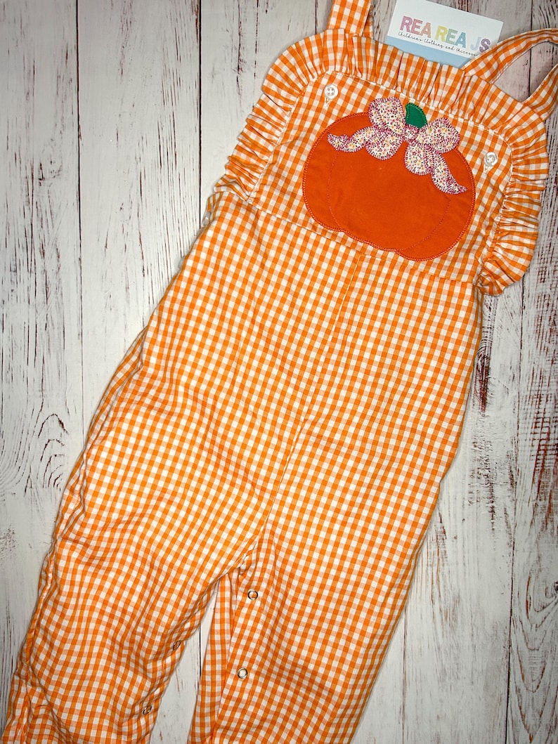 May include: Orange and white gingham overall romper with a pumpkin applique and a pink bow. The romper has ruffles on the shoulders and is made of a soft, comfortable fabric.  REA REA.S Children's Clothing and Accessories.