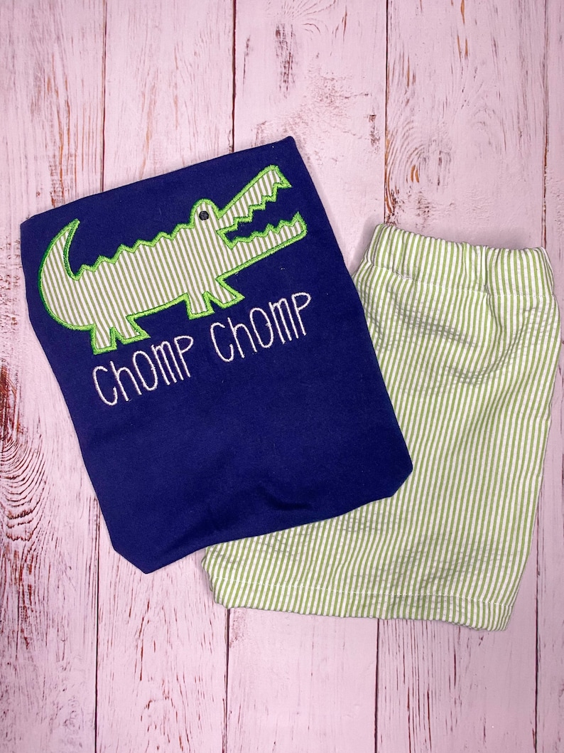 May include: A navy blue shirt with a green alligator applique and the text "CHOMP CHOMP" in white. The shirt is paired with green and white striped shorts.