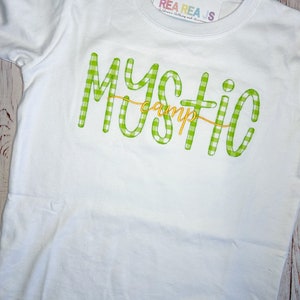 May include: White t-shirt with a green and white gingham design that says "Mystic Camp" in yellow lettering.