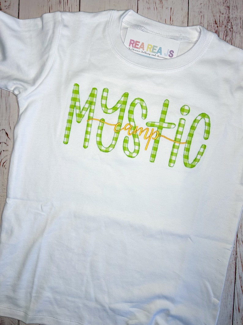 May include: White t-shirt with a green and white gingham design that says "Mystic Camp" in yellow lettering.