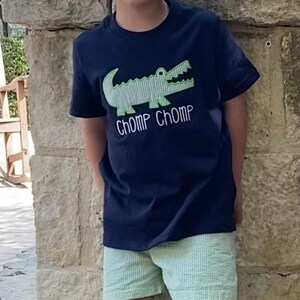 May include: A young boy wearing a navy blue t-shirt with a green alligator graphic and the text "CHOMP CHOMP". He is also wearing light green striped shorts and green and white patterned shoes.