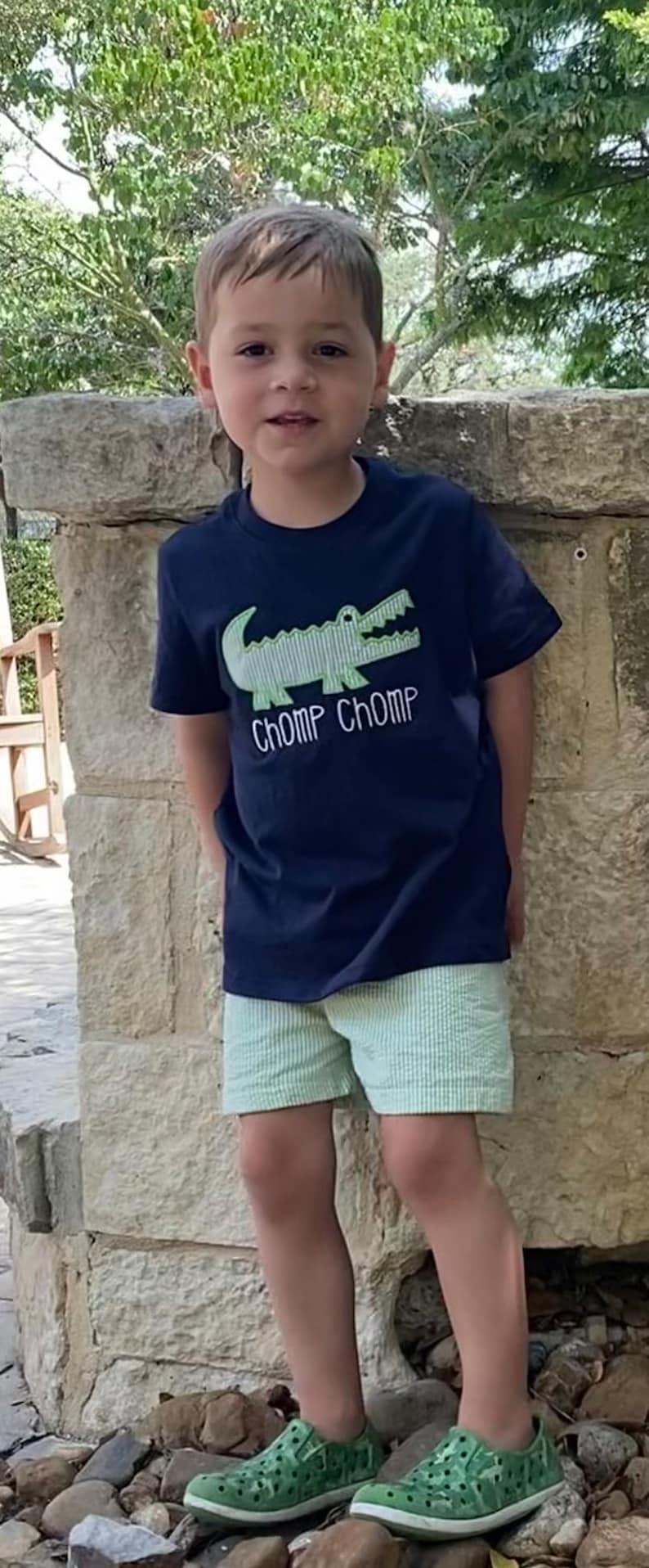 May include: A young boy wearing a navy blue t-shirt with a green alligator graphic and the text "CHOMP CHOMP". He is also wearing light green striped shorts and green and white patterned shoes.