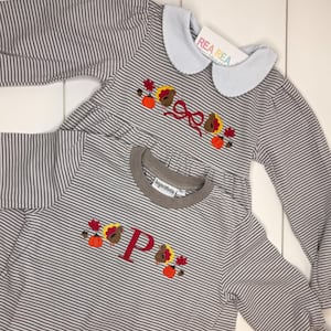 Puede incluir: Dos camisas de manga larga a rayas grises y blancas. Una tiene cuello azul claro y bordado de pavo y calabaza. La otra tiene cuello marrón y letra "P" burdeos con bordado de pavo y calabaza. De Rea Rea.