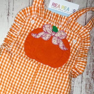 May include: Orange and white gingham overall dress with a large orange pumpkin applique with a pink bow. The dress has ruffles around the neckline and straps.