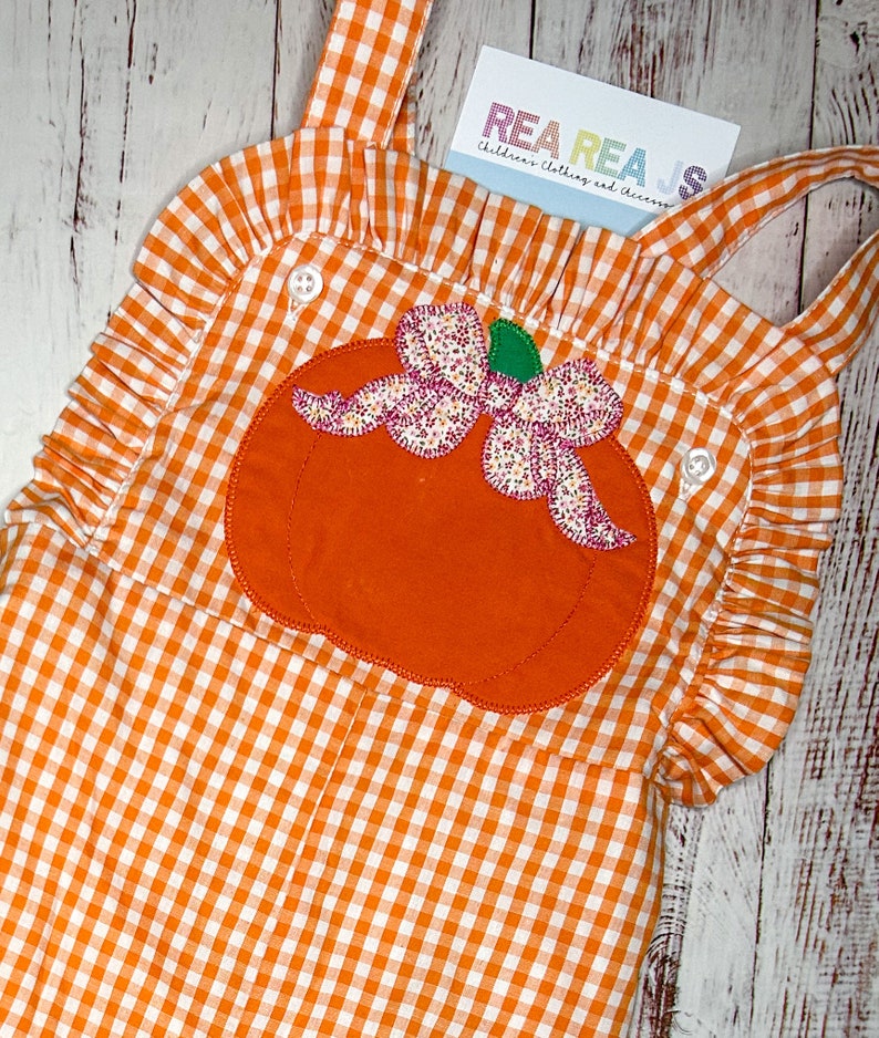 May include: Orange and white gingham overall dress with a large orange pumpkin applique with a pink bow. The dress has ruffles around the neckline and straps.