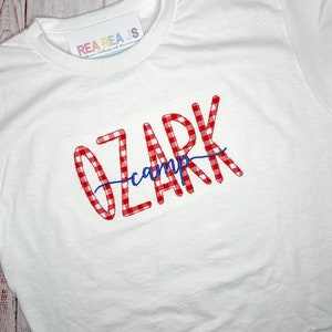 May include: White t-shirt with red and white gingham text that says "Ozark camp" in a cursive font.