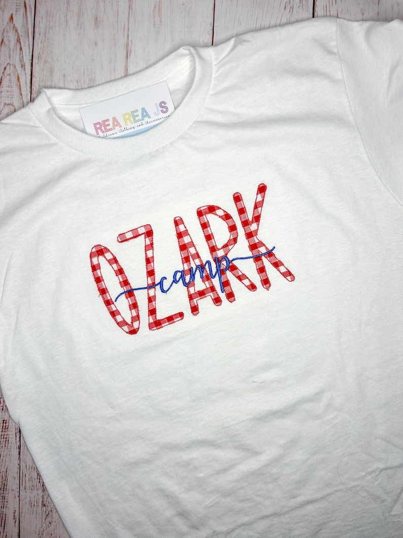 May include: White t-shirt with red and white gingham text that says "Ozark camp" in a cursive font.
