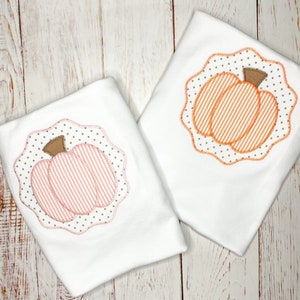 Puede incluir: Dos camisetas blancas con apliques de calabaza. Una calabaza es naranja con rayas verticales, la otra rosa con rayas horizontales. Ambas tienen tallos marrones y están enmarcadas por un borde festoneado con lunares grises.