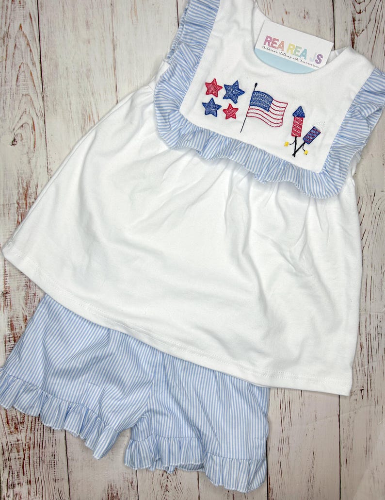 May include: A white sleeveless dress with blue and white stripes on the neckline and a red, white, and blue embroidered design of stars, a flag, and fireworks. The dress is paired with blue and white striped shorts with a ruffled hem.