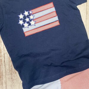 Op de afbeelding: Een marineblauw t-shirt met korte mouwen en een geborduurd vlagontwerp met witte sterren en rood-wit gestreepte stoffen accenten. Daaronder een korte broek van lichtblauw-wit gestreepte en rood-wit gestreepte stof.