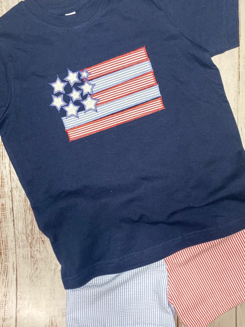 Op de afbeelding: Een marineblauw t-shirt met korte mouwen en een geborduurd vlagontwerp met witte sterren en rood-wit gestreepte stoffen accenten. Daaronder een korte broek van lichtblauw-wit gestreepte en rood-wit gestreepte stof.