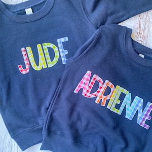 May include: Two navy blue sweatshirts with the names "JUDE" and "ADRENNE" spelled out in colorful gingham fabric.