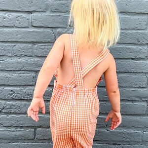May include: A young child wearing a white and orange gingham overall with a ruffled hem.