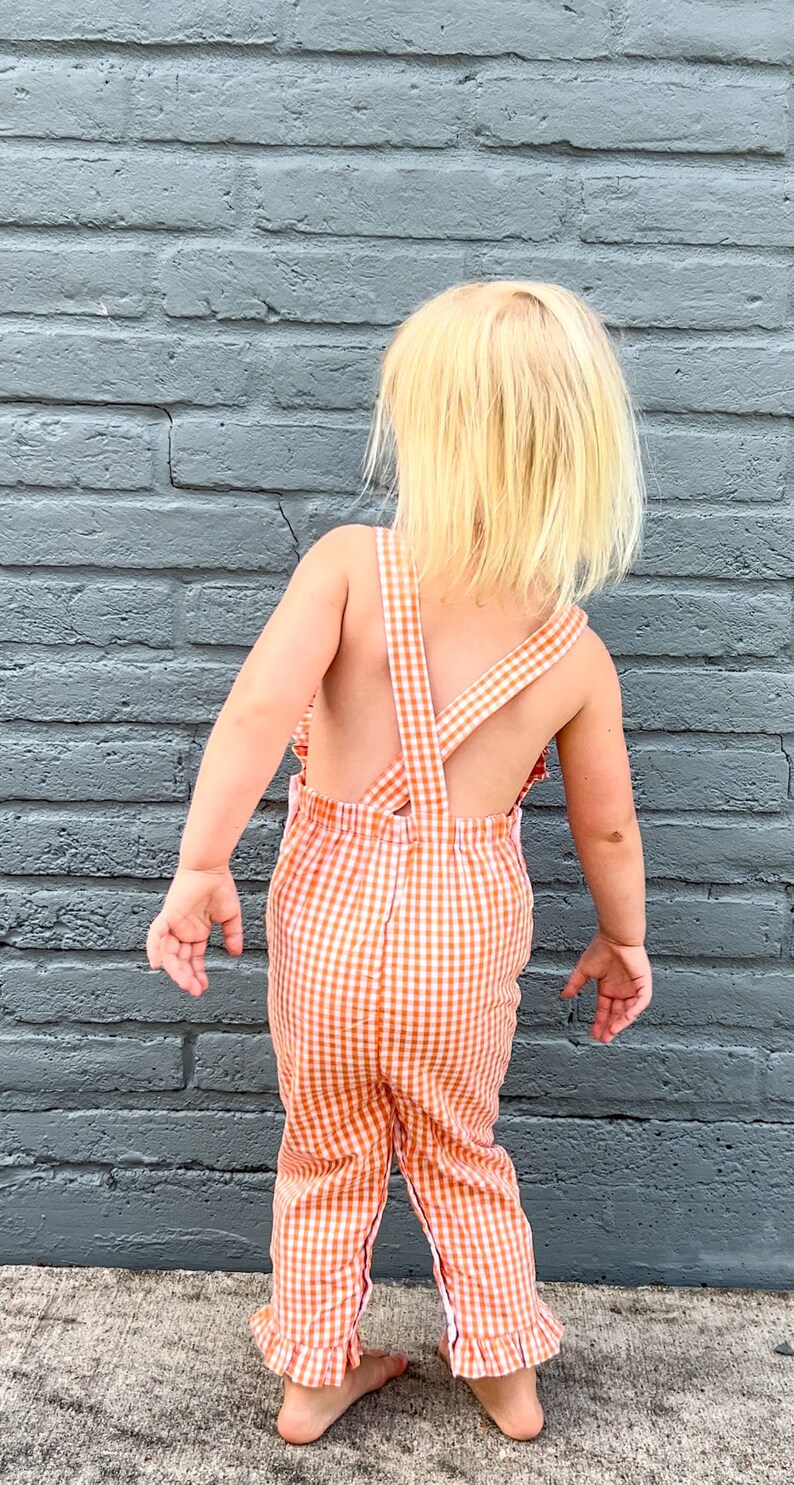 May include: A young child wearing a white and orange gingham overall with a ruffled hem.