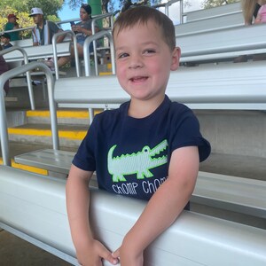 May include: A young boy wearing a navy blue t-shirt with a green alligator graphic and the text "Chomp Chomp" is smiling at the camera. He is standing in a bleacher seating area.