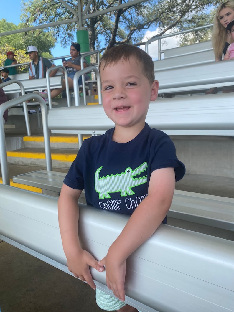 May include: A young boy wearing a navy blue t-shirt with a green alligator graphic and the text "Chomp Chomp" is smiling at the camera. He is standing in a bleacher seating area.