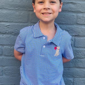 Pu&ograve; includere: Un bambino piccolo che indossa una polo a righe blu e bianche con un piccolo disegno di cane ricamato. Il cane tiene un cuore rosso.