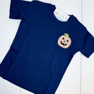 Puede incluir: Una camiseta azul marino de manga corta con un aplique de calabaza. La calabaza tiene un diseño a rayas naranja y blanco con un tallo verde y una cara tallada. La camiseta tiene una etiqueta adjunta.