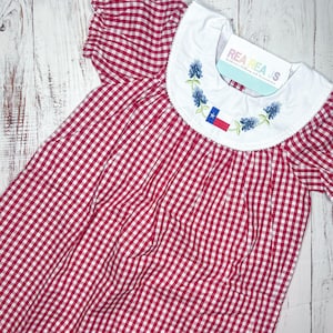 May include: Red and white gingham dress with a white collar and blue embroidered Texas bluebonnets and a Texas flag. The dress is made by Rhea's Children's Clothing and Accessories.