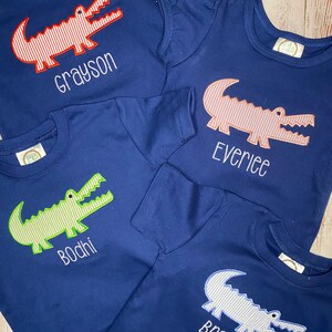 May include: Four navy blue t-shirts with alligator designs and personalised names. The alligators are red and white, pink and white, green and white, and blue and white. The names are Grayson, Everlee, Bodhi, and Bradley.